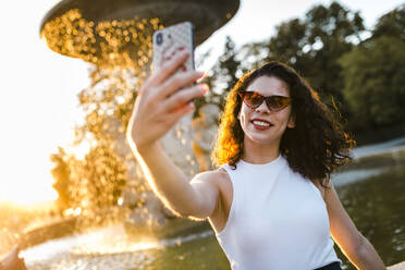 Young woman wearing sunglasses taking selfie with smart phone against fountain in park - JMPF00835