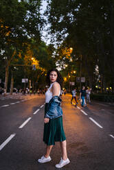 Nachdenkliche junge Frau in Jeansjacke auf der Straße vor den Bäumen in der Dämmerung - JMPF00836