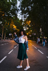 Young woman wearing denim jacket posing while standing on road in park at dusk - JMPF00837