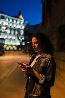 Junge Frau in Jeansjacke benutzt ihr Smartphone, während sie in der Abenddämmerung auf der Straße steht - JMPF00839