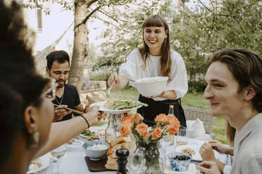 Lächelnde Frau, die einer Freundin über den Tisch hinweg Essen reicht, während einer Gartenparty - MASF21266