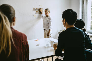 Mature businesswoman writing on adhesive note while explaining colleagues during seminar in office - MASF21290