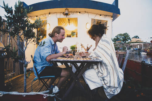Seitenansicht eines heterosexuellen Paares, das während des Sonnenuntergangs in einem Hausboot am Tisch sitzt - MASF21305