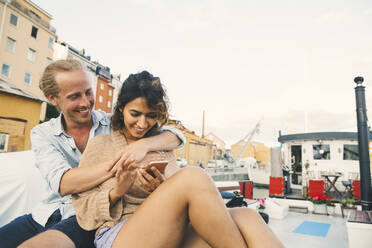 Lächelnde Freundin, die ihr Smartphone benutzt, während ihr Freund sie auf einem Hausboot umarmt - MASF21314