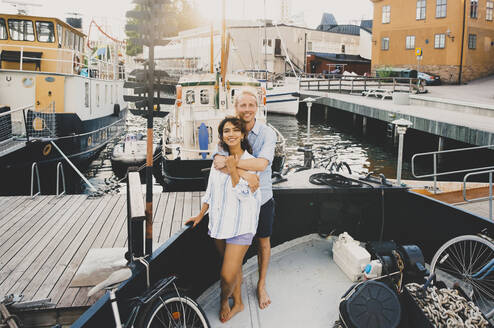 Smiling boyfriend embracing girlfriend while standing in houseboat at harbor - MASF21320