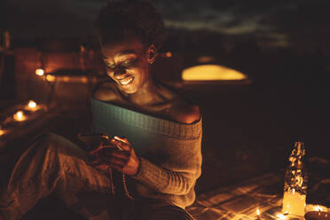 Glückliche Frau, die ein Mobiltelefon benutzt, während sie auf einer Picknickdecke auf dem Dach sitzt - MASF21341