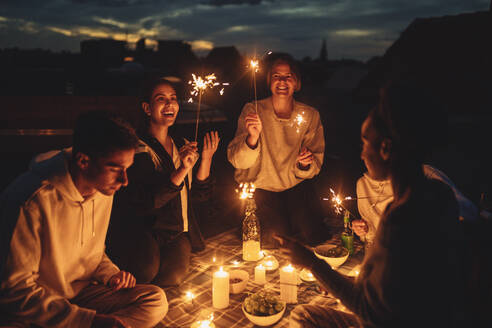 Lächelnde weibliche und männliche Freunde, die nachts auf der Terrasse eines Gebäudes einen Sprinkler zünden - MASF21349