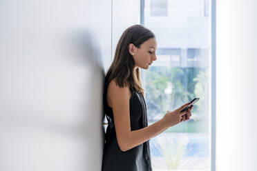 Teenager-Mädchen, das ein Mobiltelefon benutzt, während es an einer weißen Wand zu Hause steht - DLTSF01521