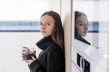 Serious teenage girl with drink standing by appliance in kitchen - DLTSF01528