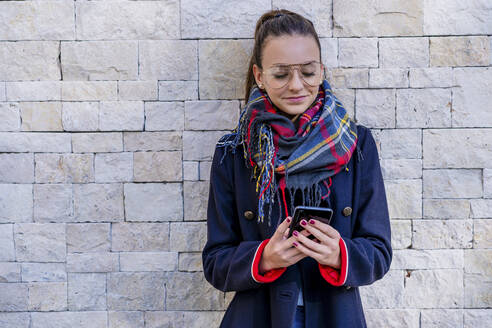 Teenage girl wearing warm clothing using mobile phone while standing against brick wall at home - DLTSF01535