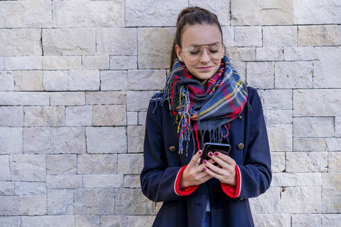 Teenager-Mädchen in warmer Kleidung, das ein Mobiltelefon benutzt, während es zu Hause an einer Mauer steht - DLTSF01535