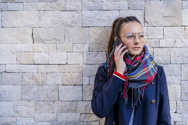 Teenager-Mädchen in warmer Kleidung im Gespräch über Smartphone gegen eine Backsteinmauer zu Hause - DLTSF01536