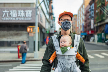 Vater mit Gesichtsmaske, der seine Tochter in der Babytrage trägt, während er in der Stadt während der Pandemie die Straße überquert - CAVF91934