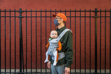 Vater mit Gesichtsmaske trägt seine süße Tochter in der Babytrage und steht während der Coronavirus-Krise am Zaun - CAVF91948