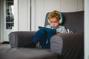Junge schmollt, spielt mit einem Tablet und trägt Kopfhörer zu Hause covid - CAVF92016