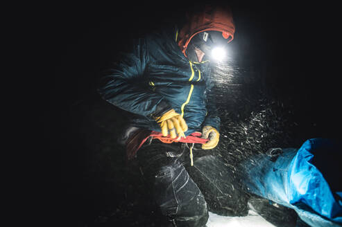 Männlicher Bergsteiger beim Anziehen einer Jacke in der Dunkelheit bei Schneetreiben - CAVF92025