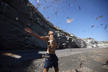 Shirtless man with arms outstretched standing under leaves falling on him at beach - AXHF00099