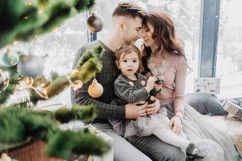 Young family with a daughter in festive outfits with a garland near the Christmas tree on New Years Eve - CAVF92081