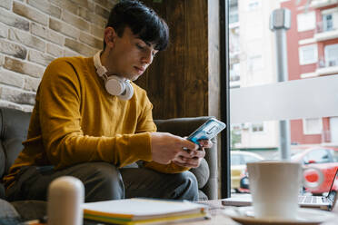 Junger Mann mit Kopfhörern, der in einem Café sitzt und ein Mobiltelefon benutzt - EGAF01461