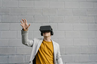 Junger Mann mit Virtual-Reality-Headset, der seine Hand ausstreckt, während er an der Wand steht - EGAF01465