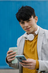 Lächelnder Mann mit Kaffeetasse, der ein Mobiltelefon benutzt, während er an einer blauen Wand steht - EGAF01472