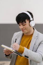 Smiling man wearing headphones writing in book while standing against wall - EGAF01474
