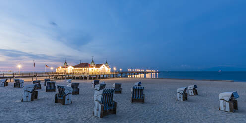 Germany, Mecklenburg-Western Pomerania, Heringsdorf, Hooded beach chairs on empty beach at dusk with illuminated Ahlbeck Pier in background - WDF06495
