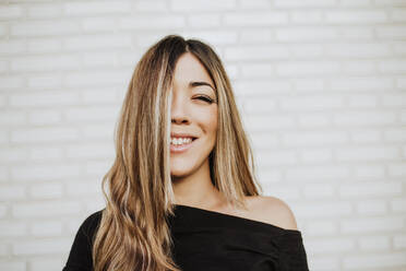 Close-up of smiling young woman covering face with hair against white wall - MIMFF00464