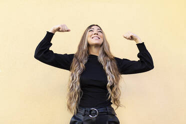 Junge Frau mit langen Haaren, die ihre Muskeln anspannt, während sie vor einem beigen Hintergrund nach oben schaut - MIMFF00468