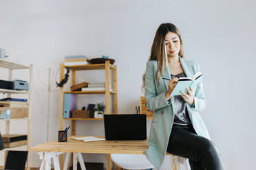 Smiling businesswoman writing in diary while sitting on desk against wall - MIMFF00481