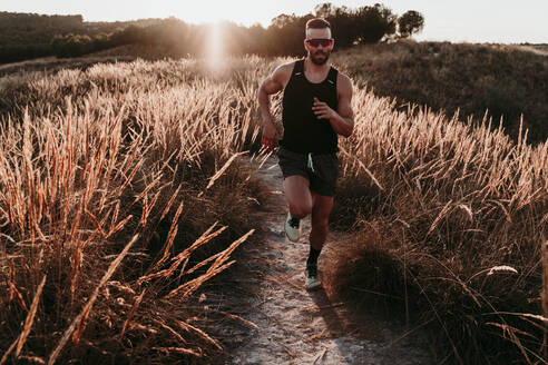 Junger männlicher Sportler, der bei Sonnenuntergang auf einem Berg in Spanien im getrockneten Gras läuft - EBBF02281