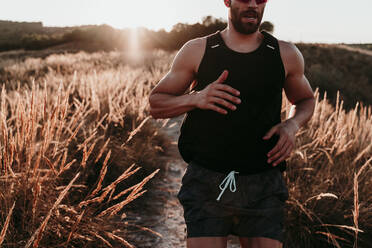Männlicher Sportler, der bei Sonnenuntergang durch getrocknetes Grasfeld läuft - EBBF02282