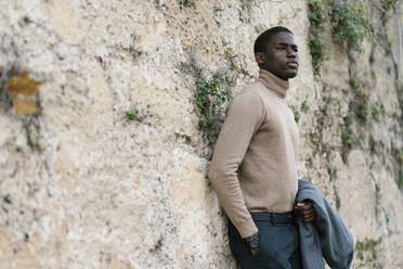 Fashionable man with hands in pockets holding jacket while leaning on wall - EGAF01492