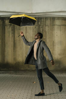 Junger Mann mit Jacke, der einen Regenschirm hält, während er auf einem Fußweg geht - EGAF01504