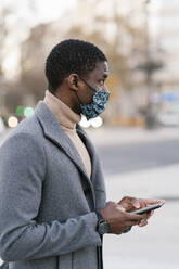 Mann mit Gesichtsschutzmaske, der im Freien stehend ein Mobiltelefon benutzt - EGAF01510