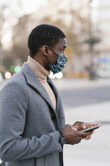 Man wearing protective face mask using mobile phone while standing outdoors - EGAF01510