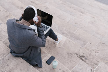 Junger Mann mit Kopfhörern, der einen Laptop benutzt, während er auf einer Treppe sitzt - EGAF01511