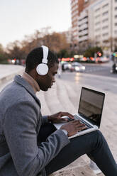 Junger Mann mit Kopfhörern, der in der Stadt sitzend am Laptop arbeitet - EGAF01517