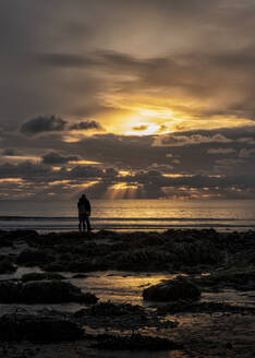 Silhouette eines Paares, das am Strand von Manorbier in Pembrokeshire, Großbritannien, steht und die Aussicht betrachtet - ALRF01796