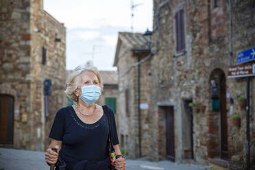 Ältere Frau mit Gesichtsschutz, die einen Wanderstock hält, während sie in einer italienischen Stadt steht - MAMF01562