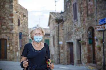 Ältere Frau mit Gesichtsschutz, die einen Wanderstock hält, während sie in einer italienischen Stadt steht - MAMF01563