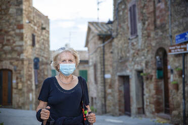 Senior woman wearing protective face mask holding hiking pole while standing in Italian city - MAMF01563