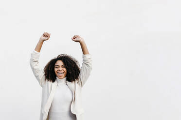 Cheerful afro woman with arms raised screaming while standing against white wall - TCEF01441