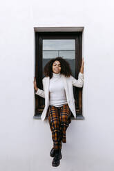Afro junge Frau lächelnd auf der Fensterbank sitzend - TCEF01445