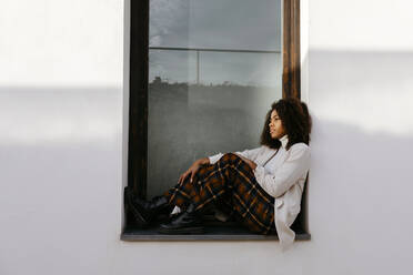Thoughtful woman with afro hair sitting on window sill of building - TCEF01447