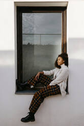 Thoughtful afro young woman sitting on window sill - TCEF01448