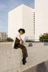 Carefree young woman with afro hair sitting on retaining wall against buildings in city - TCEF01451