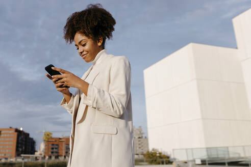 Smiling young woman with afro hair using smart phone while standing against sky in city - TCEF01454