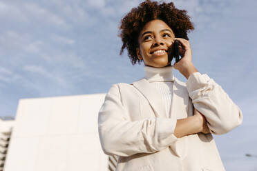Junge Frau in weißem Mantel, die über ein Mobiltelefon spricht, während sie vor dem Himmel steht - TCEF01455