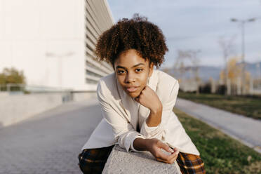 Selbstbewusste junge Frau mit Afro-Haar sitzt auf einer Stützmauer in der Stadt - TCEF01456